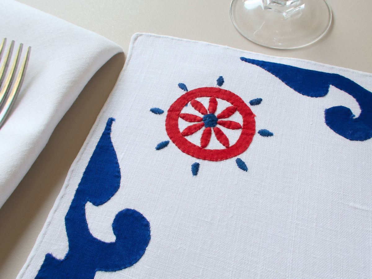 Close view of vintage Italian nautical embroidery and appliqué ship’s wheel and wave motifs in navy and red on white linen.