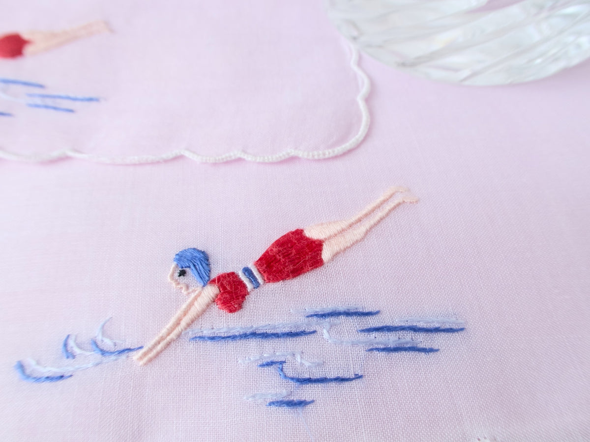 Close view of hand embroidered retro diver on vintage cotton cocktail napkin, red swimsuit, blue water detail.​​​​​​​​​​​​​​​​