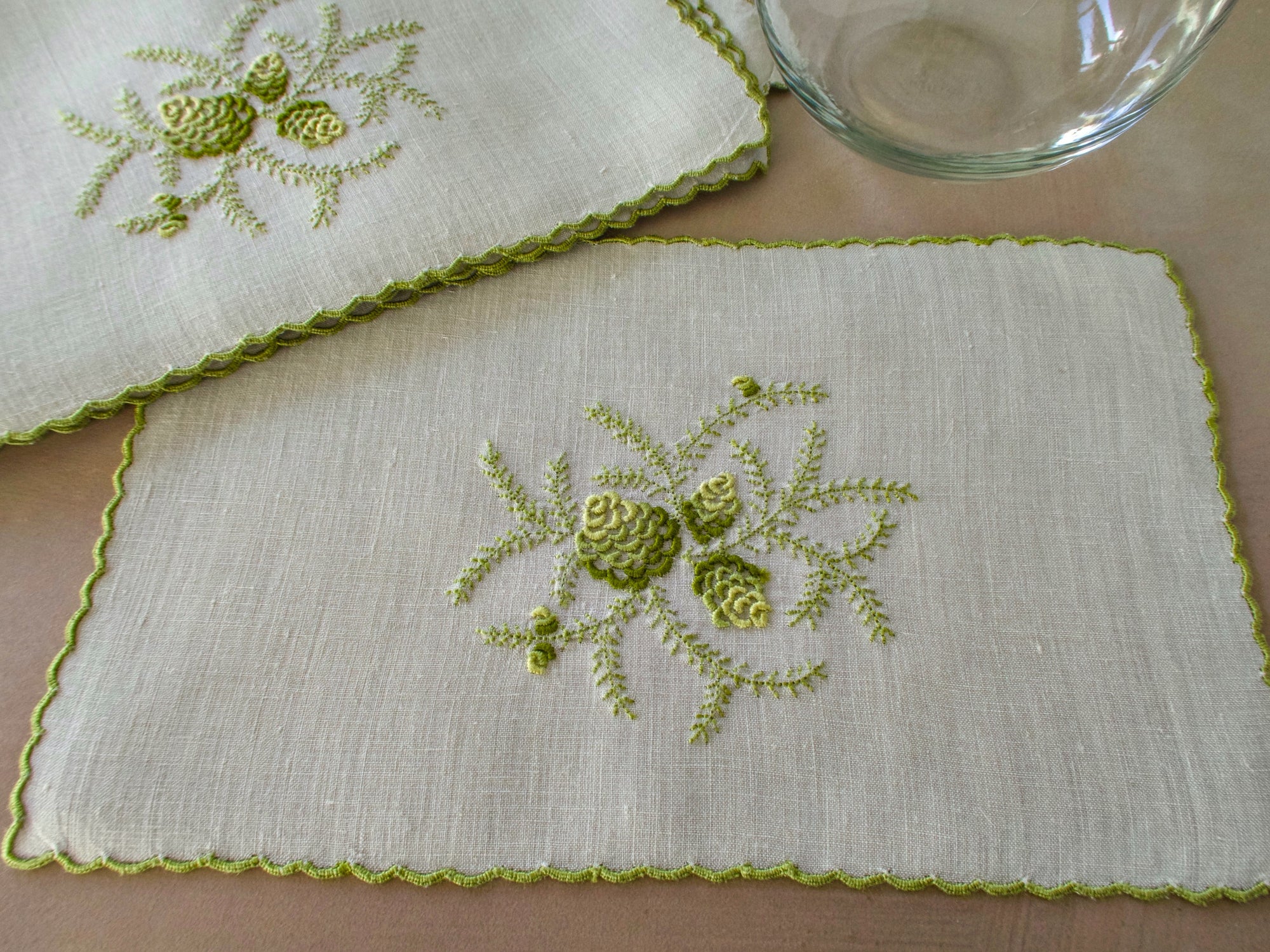 Green embroidered pinecone motif on linen cocktail napkin, scalloped edge.