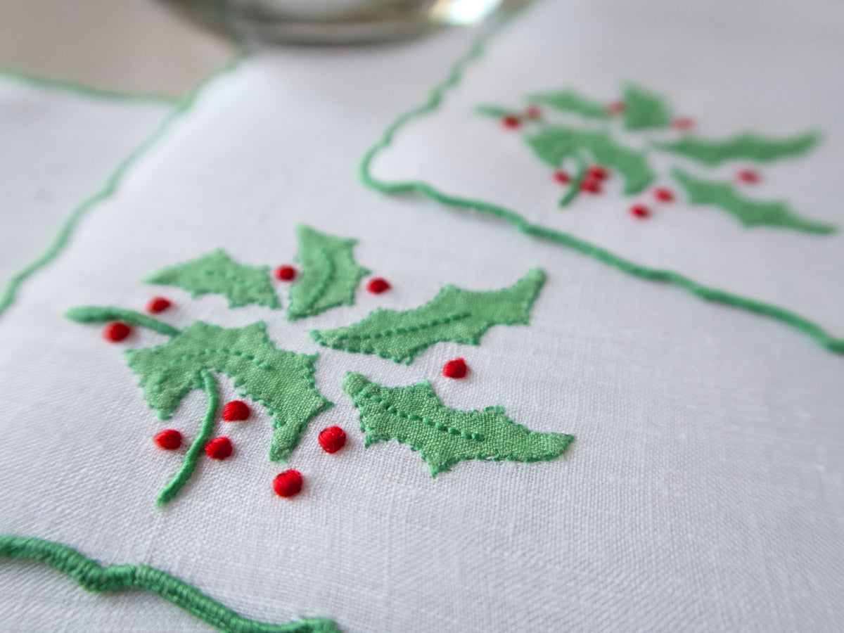 Close-up of white linen cocktail napkin with green holly leaves and red berry embroidery on a scalloped edge.