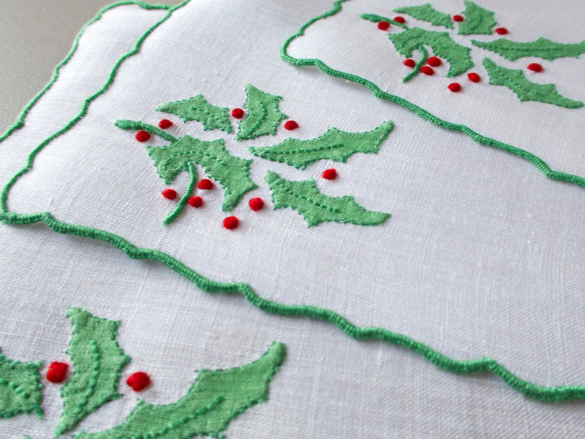 Three white linen cocktail napkins layered and angled, showing scalloped edges and embroidered holly leaves and red berries.
