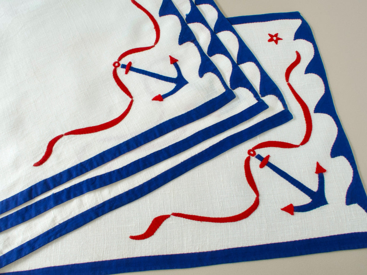Vintage nautical linen placemats stacked and fanned, showing blue anchor, red stars and scalloped blue border design.​​​​​​​​​​​​​​​​