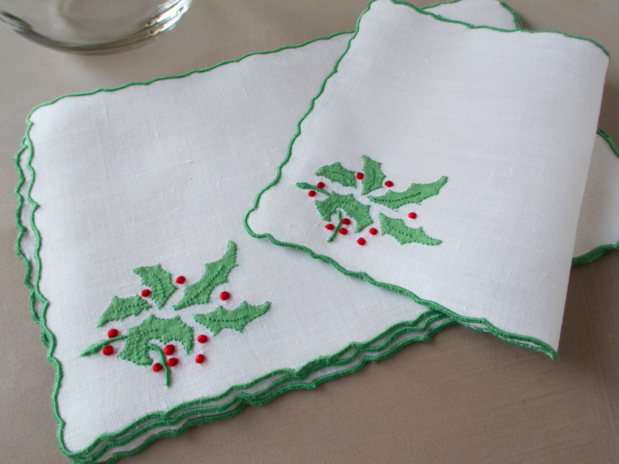 White linen cocktail napkin folded diagonally, showing scalloped border and embroidered holly leaves and red berries along one edge.