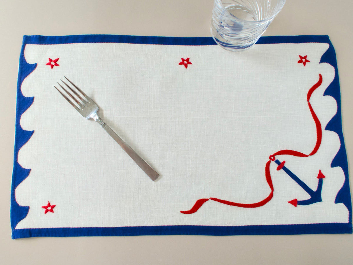 Close view of blue anchor with red ribbon and red stars on vintage white linen placemat, scalloped blue border detail.