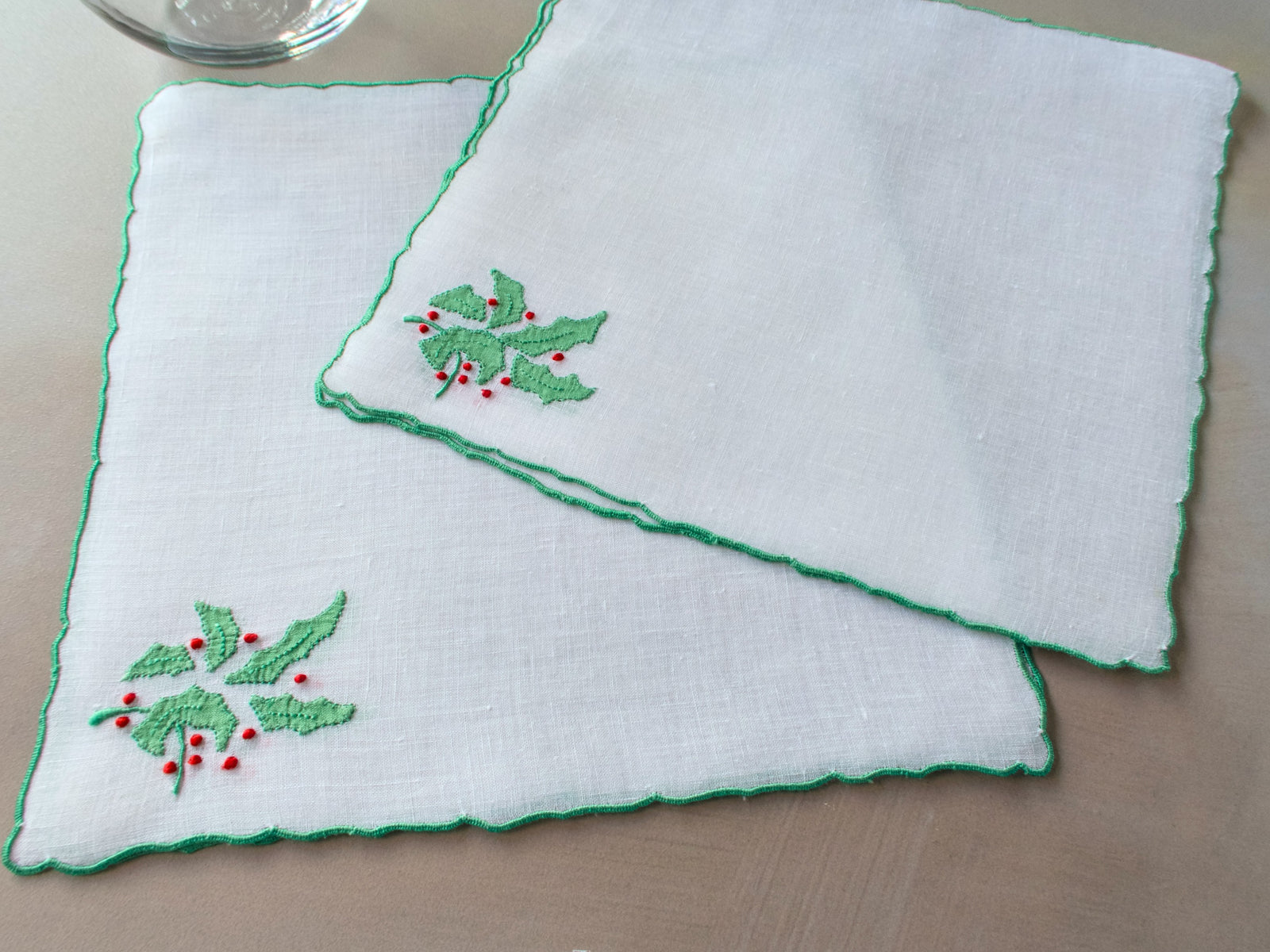 Two white square linen cocktail napkins layered and angled, showing scalloped edges and embroidered holly sprigs.