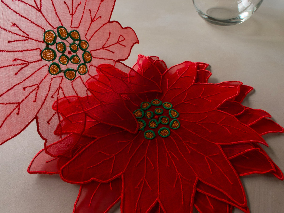Stack of red and pink poinsettia-shaped organdy cocktail napkins, layered to show petal edges and embroidered centers, one held up to show sheer fabric and stitching.
