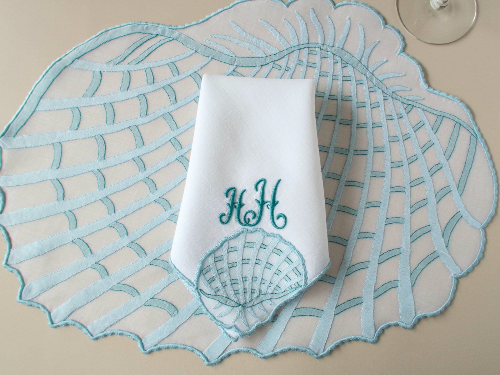 Folded monogrammed linen napkin with HH in deep teal, shell motif at corner, on beige background — Maison Léron