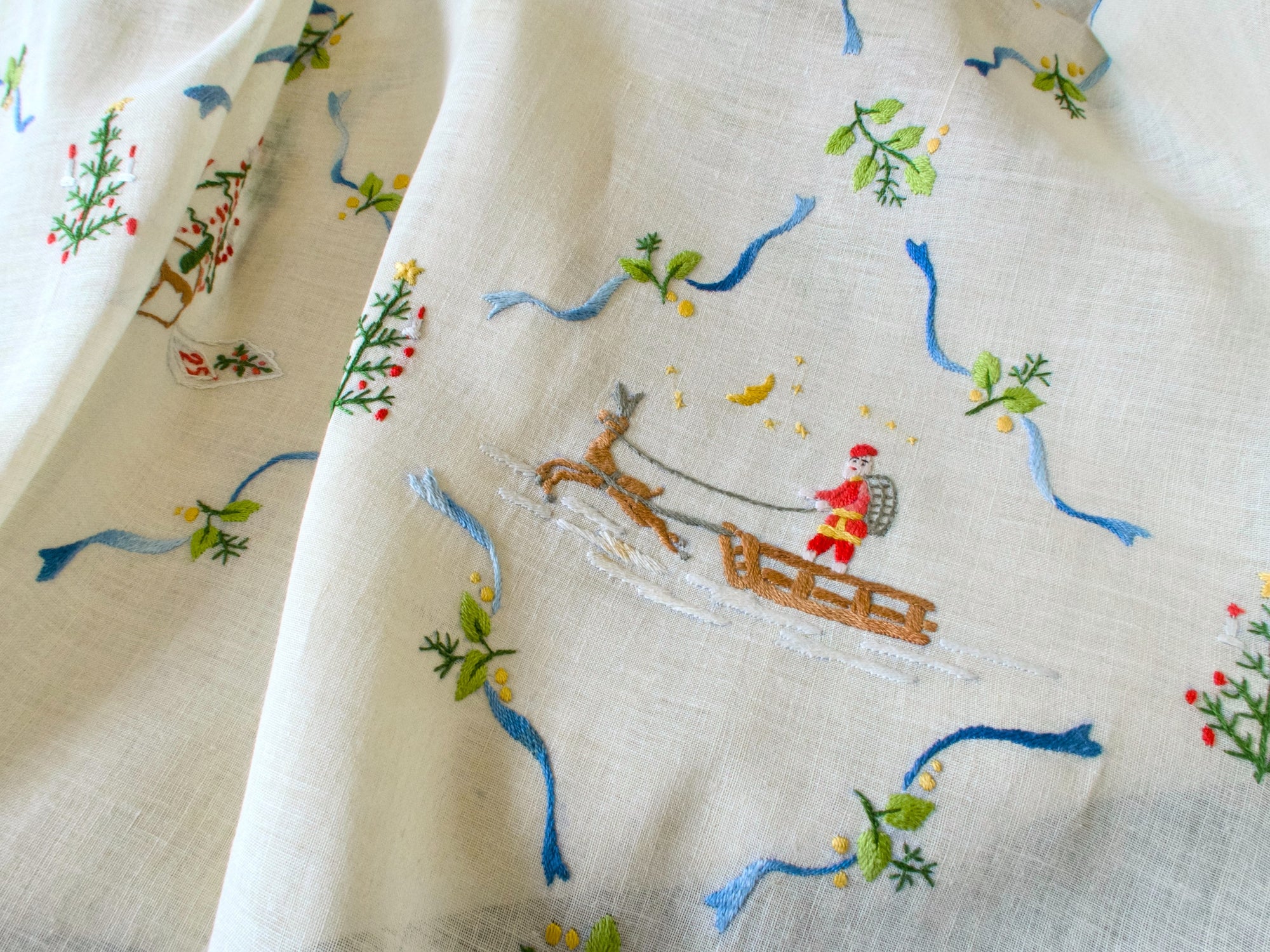 Vintage linen Christmas tablecloth with embroidered Santa and holiday motifs, detail shot showing Santa in red sled with gifts and greenery.