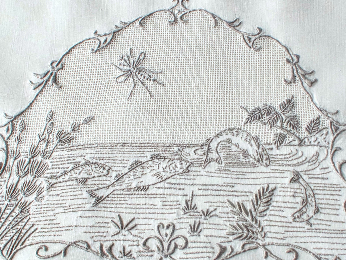 Closeup of a vintage Italian hand embroidered guest towel, fish swimming among reeds and water plants beneath open mesh sky with dragonfly, scrolling floral border.