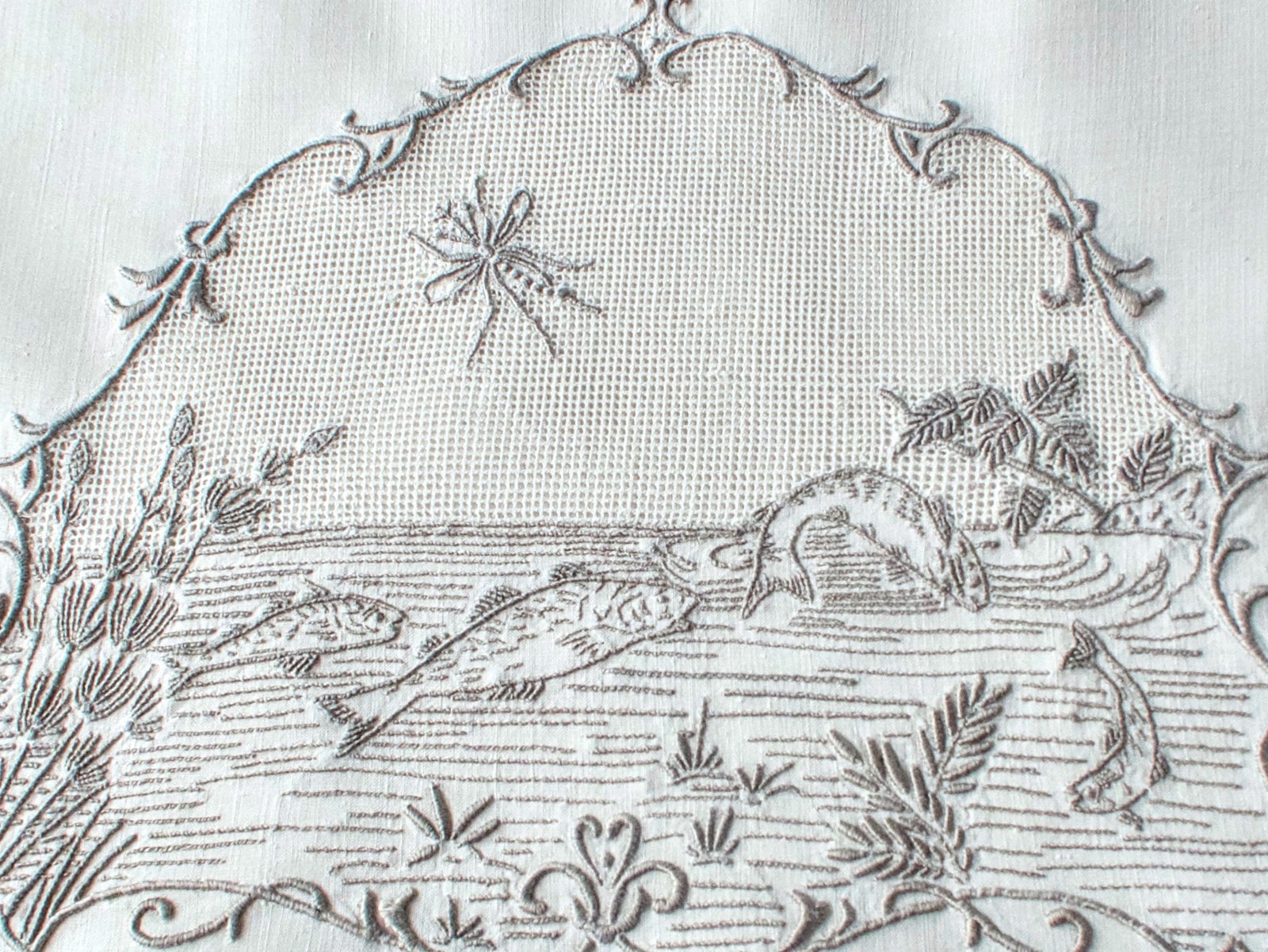 Closeup of a vintage Italian hand embroidered guest towel, fish swimming among reeds and water plants beneath open mesh sky with dragonfly, scrolling floral border.
