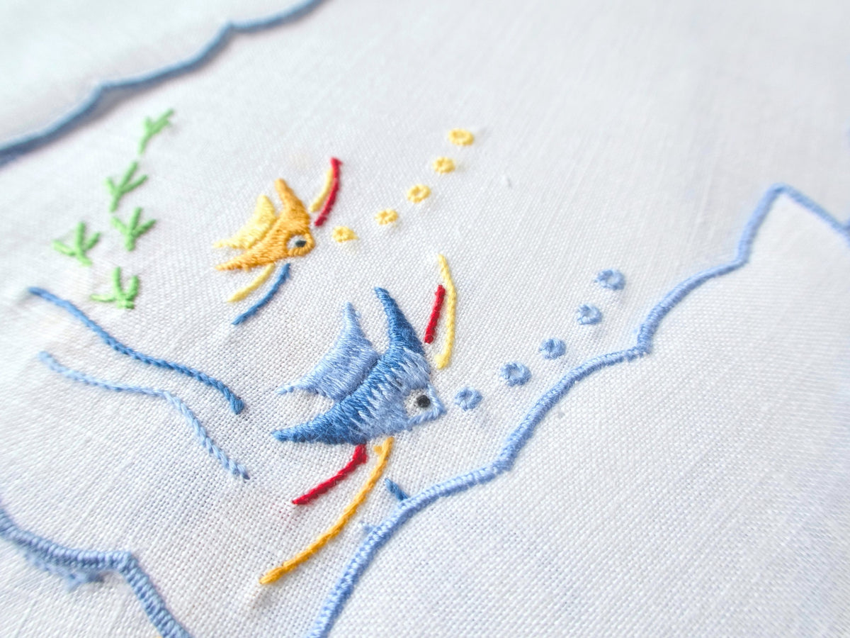 Close view of yellow and blue angelfish hand embroidery with bubbles and seaweed on vintage white linen cocktail napkin, showing small hole at edge.​​​​​​​​​​​​​​