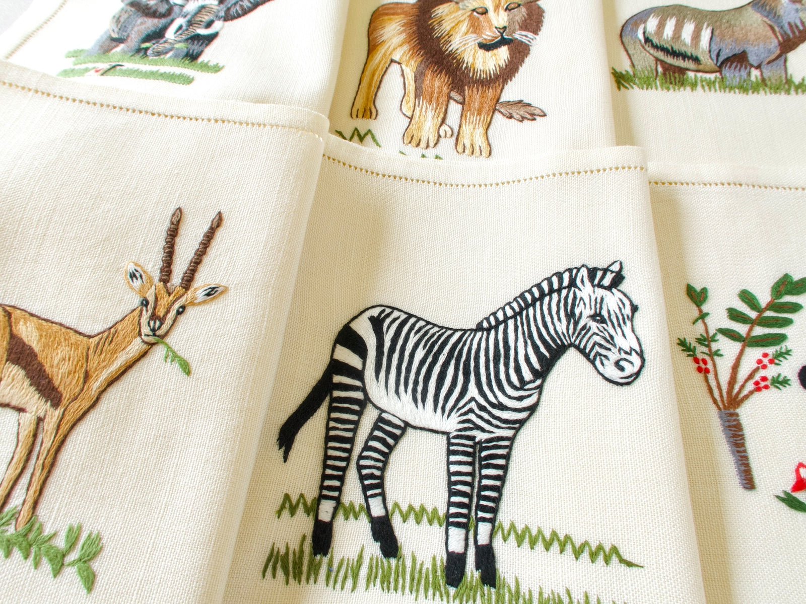 Vintage hand embroidered linen placemats fanned out showing gazelle, zebra and elephant with entwined trunks on cream linen