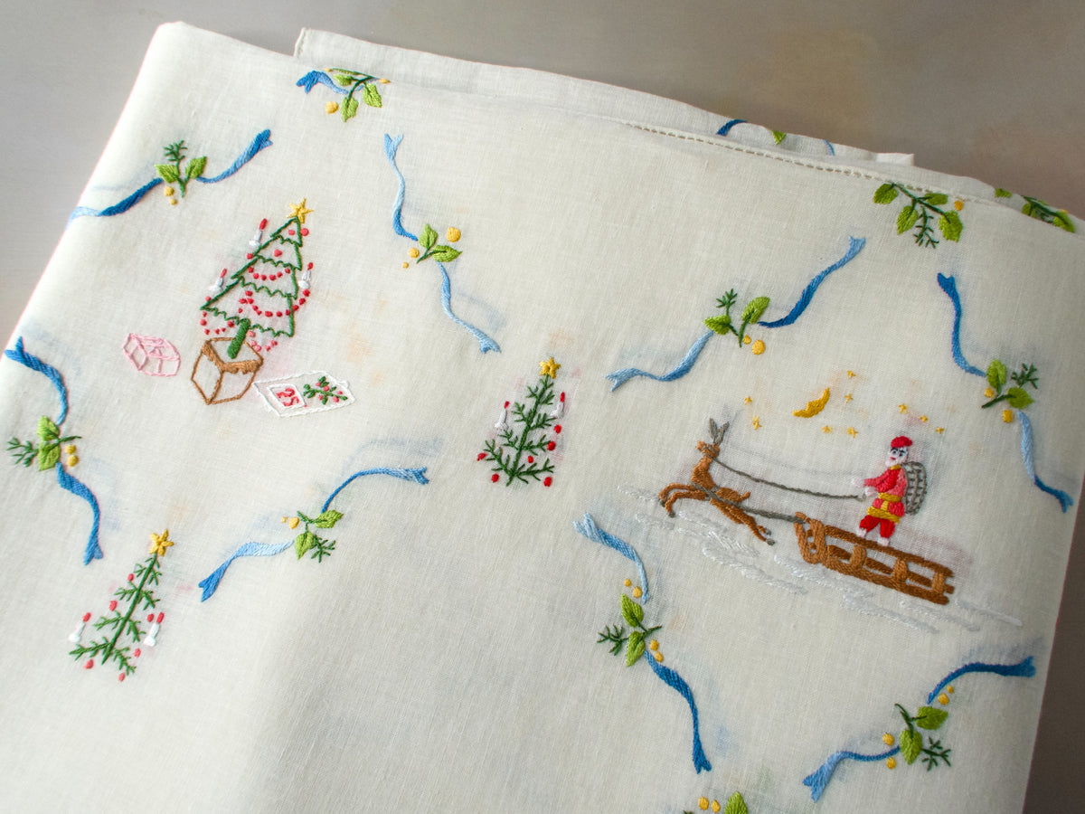 Medium view of embroidered Christmas motifs on linen tablecloth, including Santa, trees, stars, and cottages grouped together.