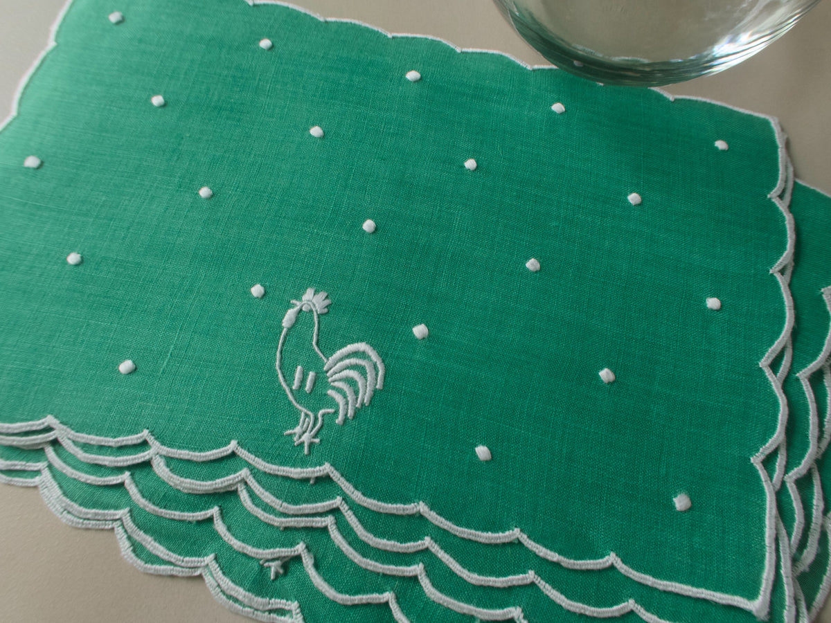 Green linen cocktail napkin with white embroidered rooster, snowy dots and scalloped border, shown flat.