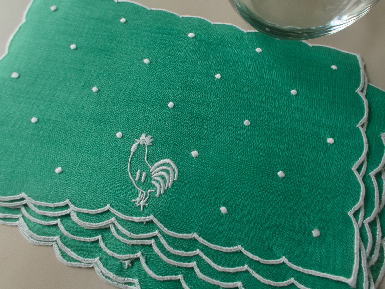 Green linen cocktail napkin with white embroidered rooster, snowy dots and scalloped border, shown flat.