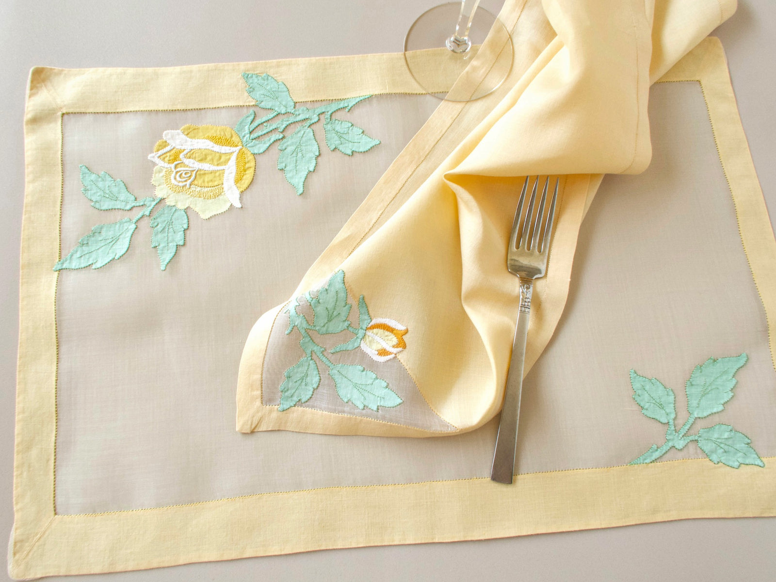 Vintage Madeira organdy placemat with folded linen napkin on top, yellow rose and sage green leaf appliqué, fork and wine glass alongside