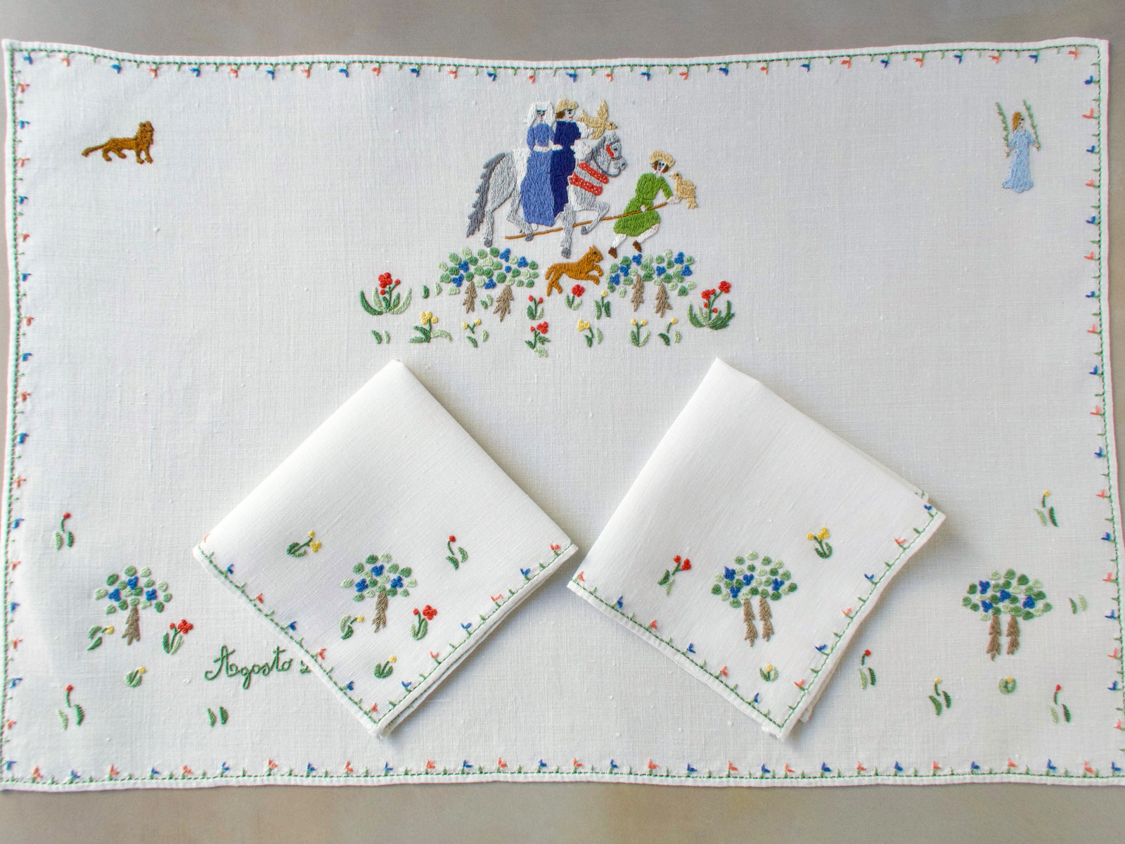 Overhead view of the August Rapisardi breakfast set with centerpiece and two napkins arranged on top, all embroidered with medieval figures and floral borders.