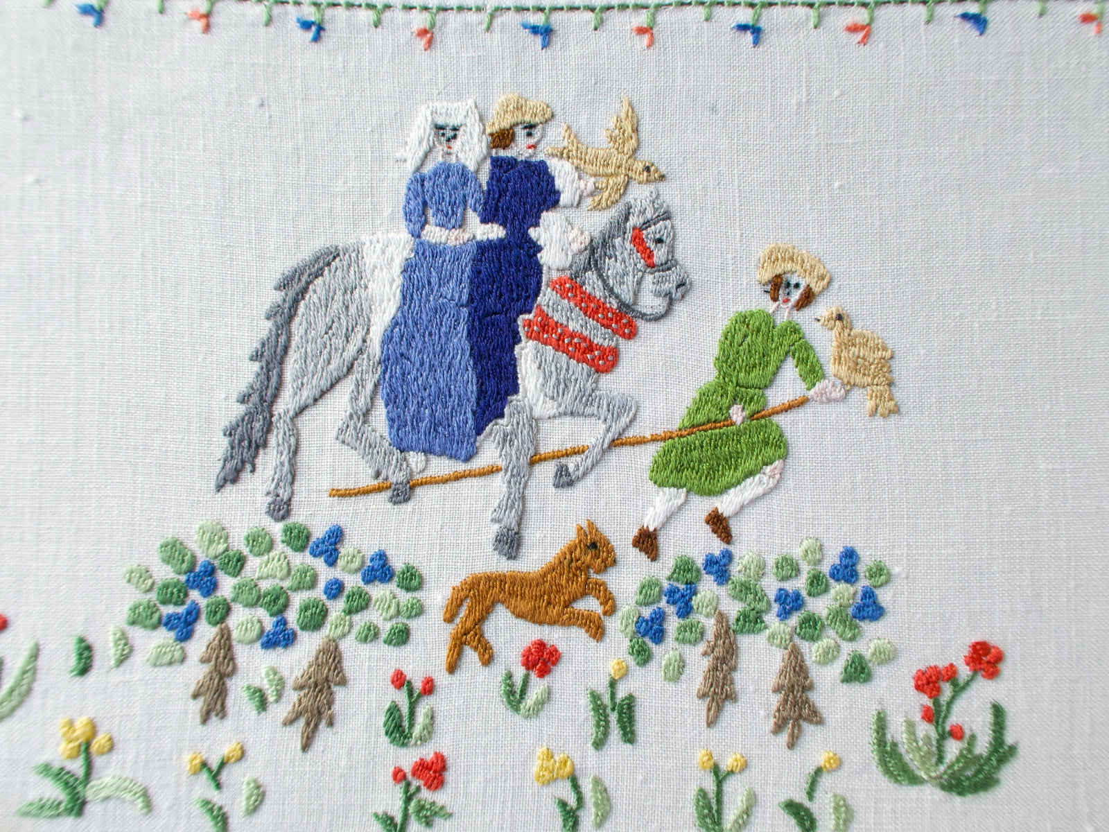 Centerpiece showing two noble riders on horseback with a dog running beside them, surrounded by raised floral embroidery in blue, green, and red.