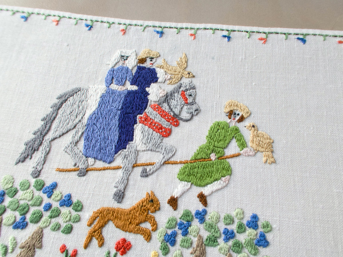 Angled close-up of the two riders on horseback in blue and green garments, with detailed shaded embroidery and a dog at their feet.