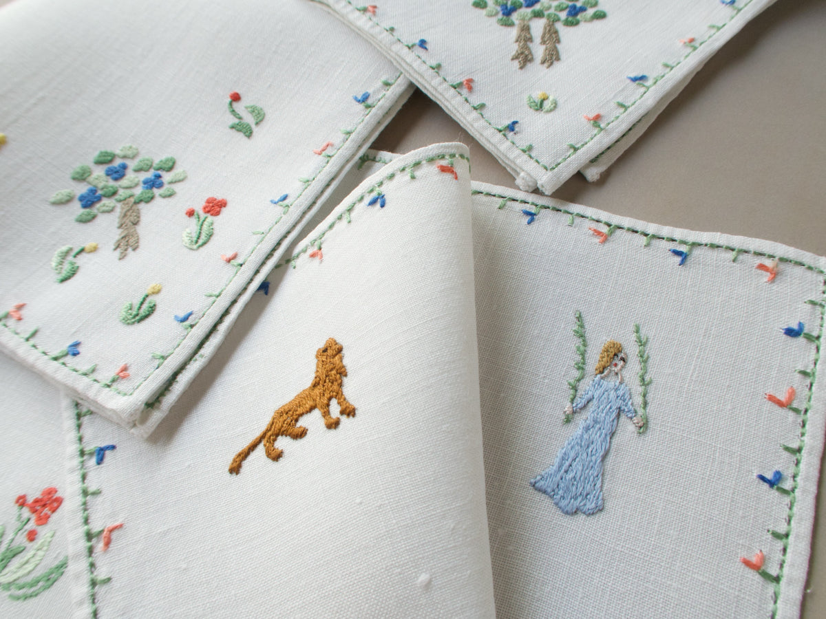 Corner of the centerpiece showing a small embroidered lion and a woman in blue near the floral border, with napkins layered above.