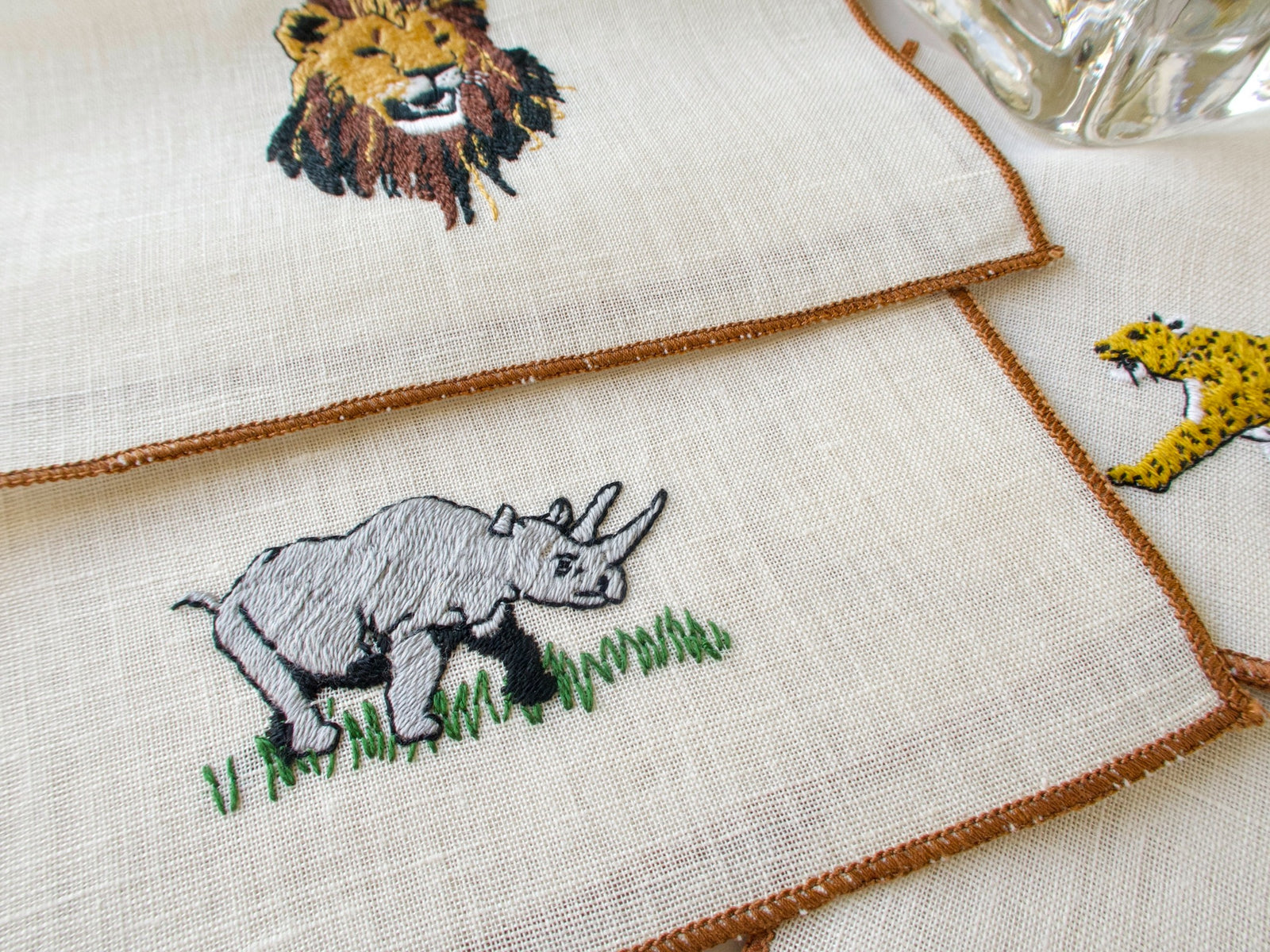 Two vintage cocktail napkins showing embroidered rhino and cheetah designs on ecru linen with brown border