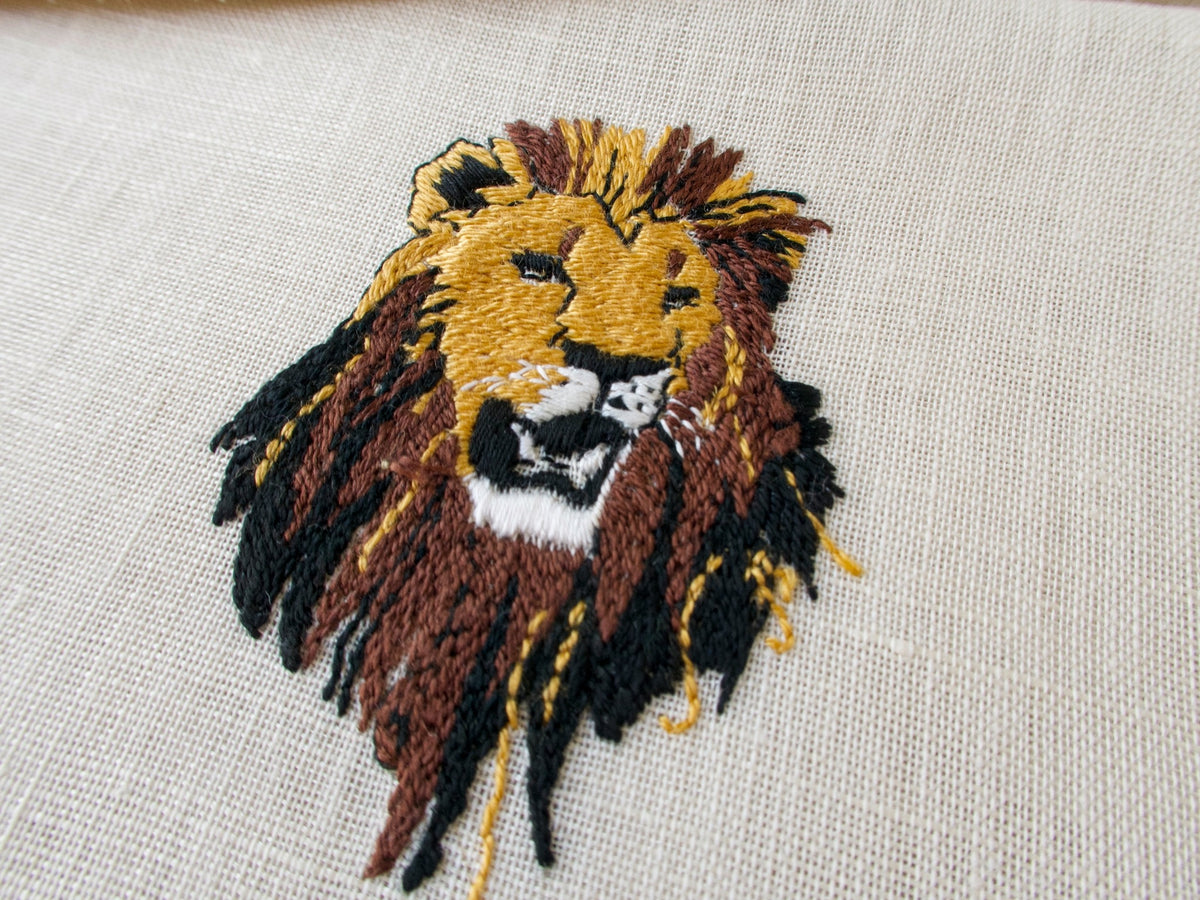 Single vintage cocktail napkin showing detailed lion head embroidery, semi-sheer ecru linen with brown border​​​​​​​​​​​​​​​​