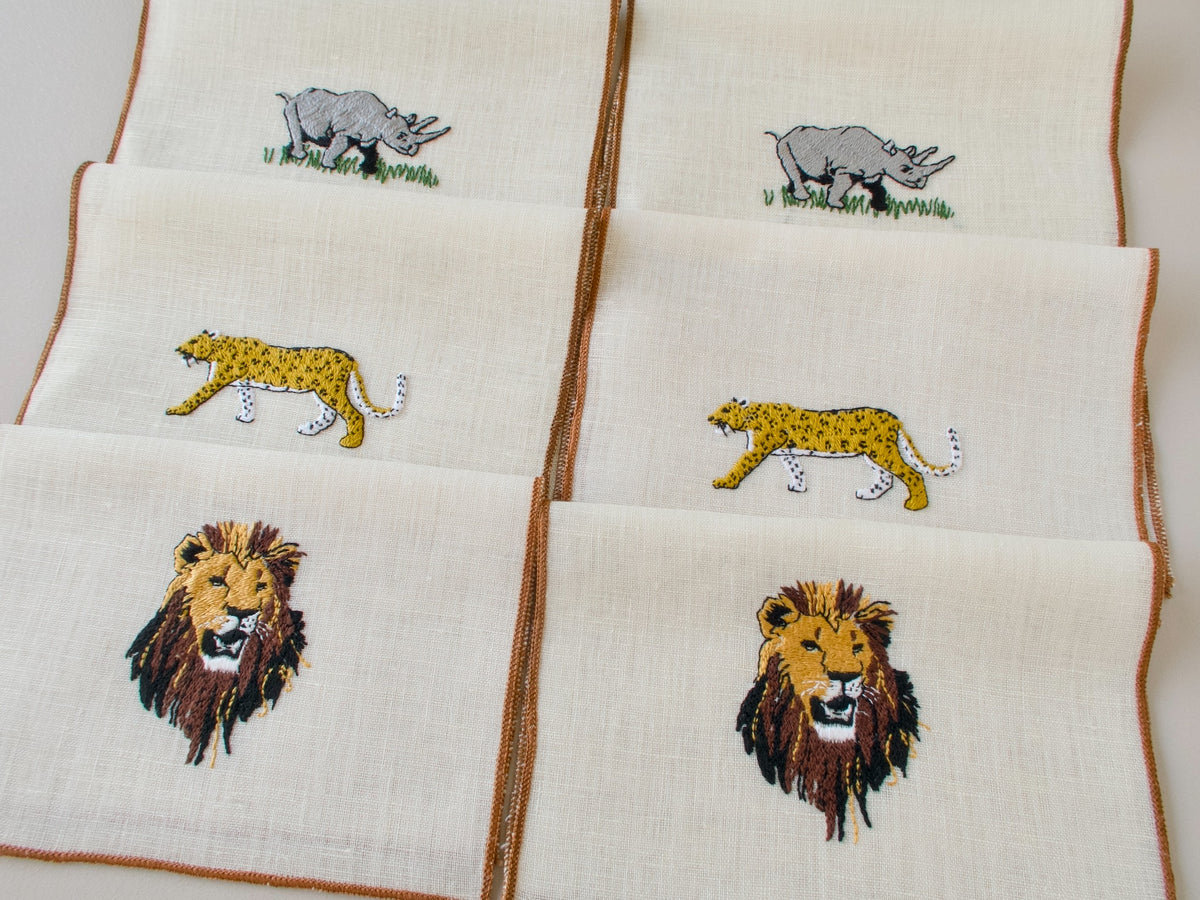 Six vintage cocktail napkins laid flat showing lion, rhino and cheetah embroidery designs