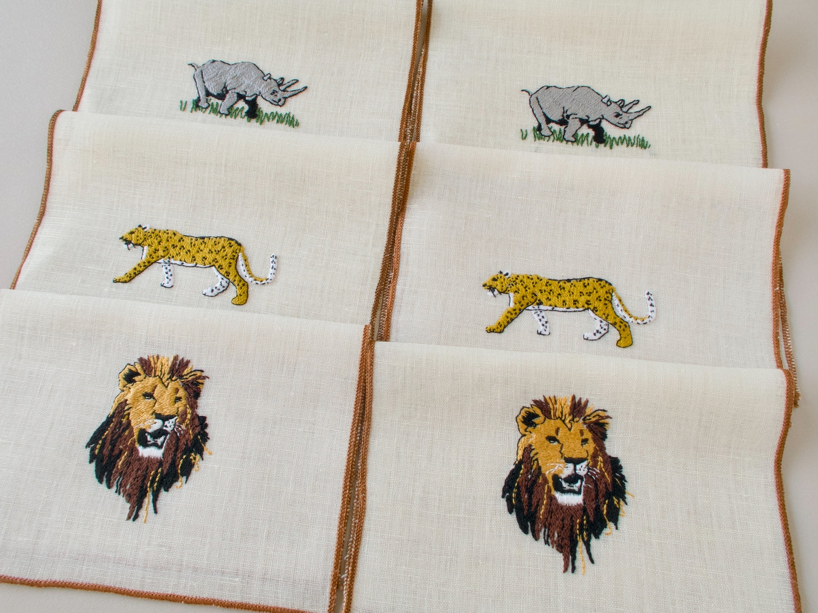Six vintage cocktail napkins laid flat showing lion, rhino and cheetah embroidery designs