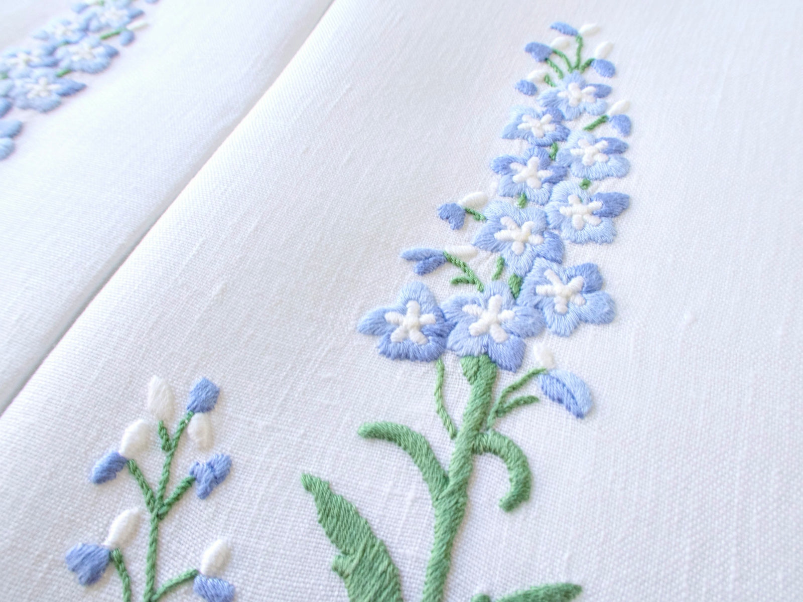Detail of raised hand embroidery on white Madeira linen, showing periwinkle blue delphinium blooms with white centers in dimensional handwork