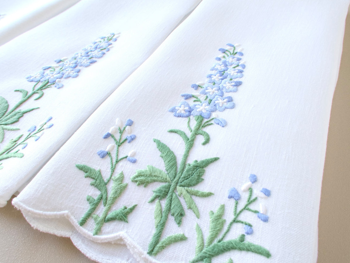 White Madeira linen guest towel folded to show the hand embroidered, dimensional delphinium design, with dense green foliage 