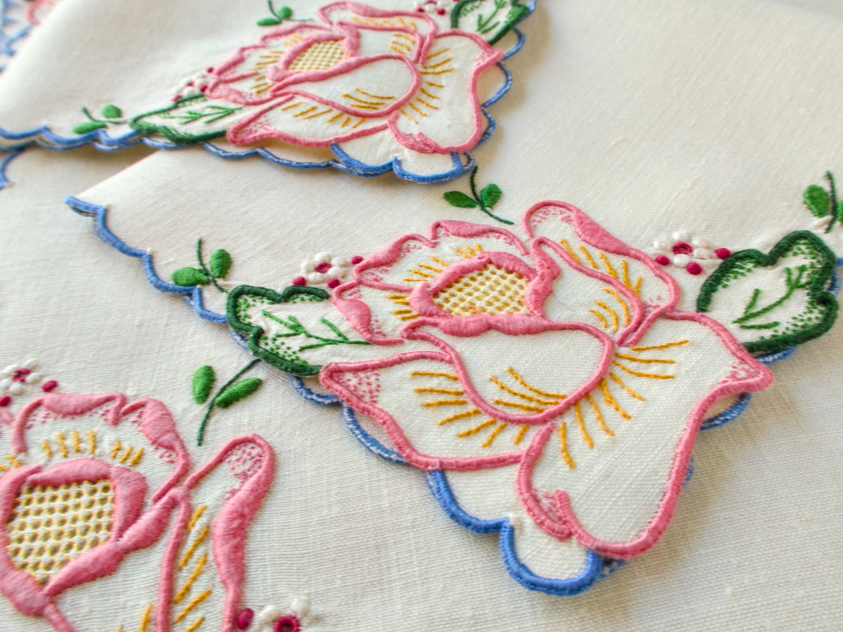 Close-up of vintage Madeira tea napkin showing large hand embroidered pink rose with drawnwork center, gold stamens and green embroidered leaves on creamy linen