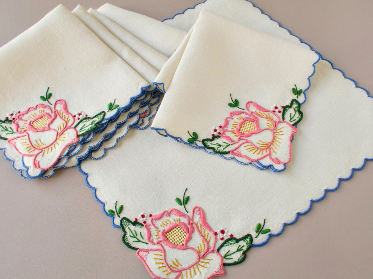Six vintage Madeira pink rose tea napkins fanned out showing full set with blue scalloped border and pink rose embroidery on white linen