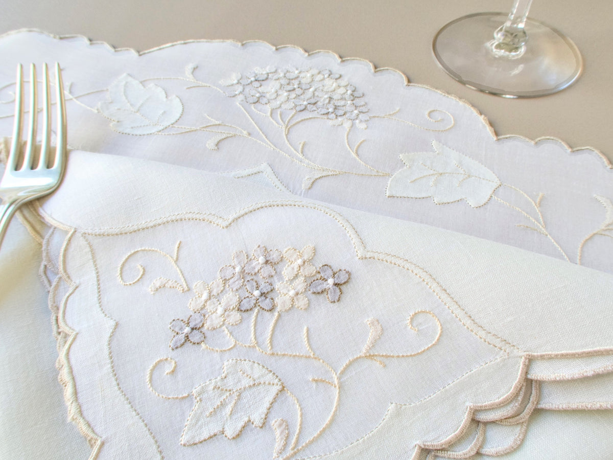 Close-up of vintage Madeira hydrangea napkins and placemat showing embroidery detail with fork and wine glass in background​​​​​​​​​​​​​​​​