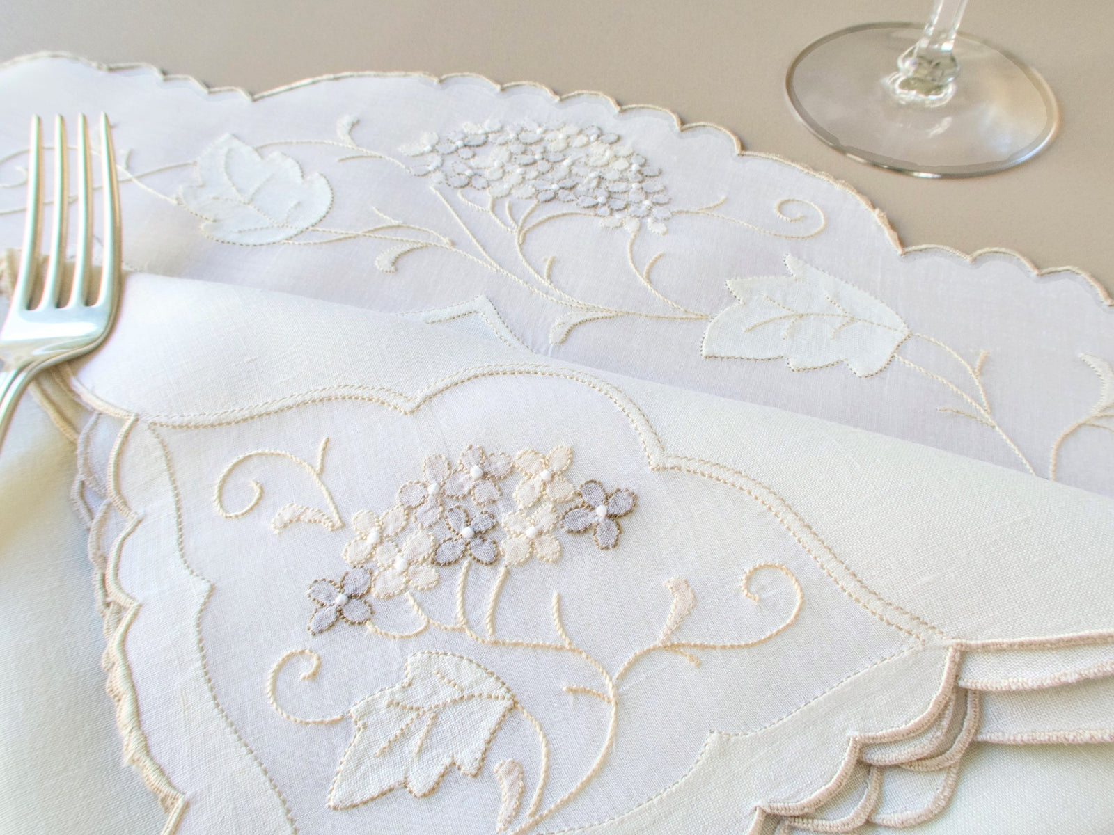 Close-up of vintage Madeira hydrangea napkins and placemat showing embroidery detail with fork and wine glass in background​​​​​​​​​​​​​​​​