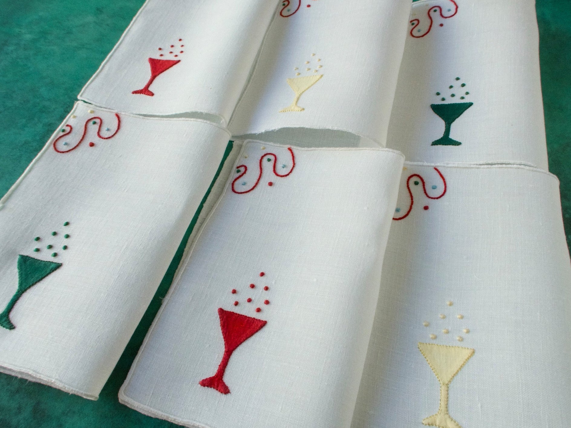 White linen cocktail napkins embroidered with champagne glasses in red, green and yellow, arranged in a group.