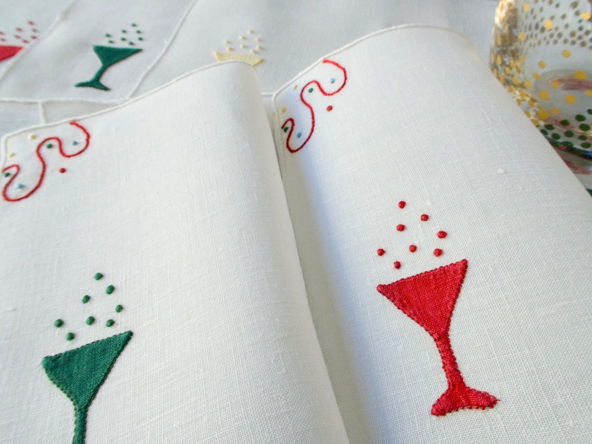 White linen cocktail napkins embroidered with red, green and yellow champagne glasses and stitched bubbles, shown folded and overlapping.