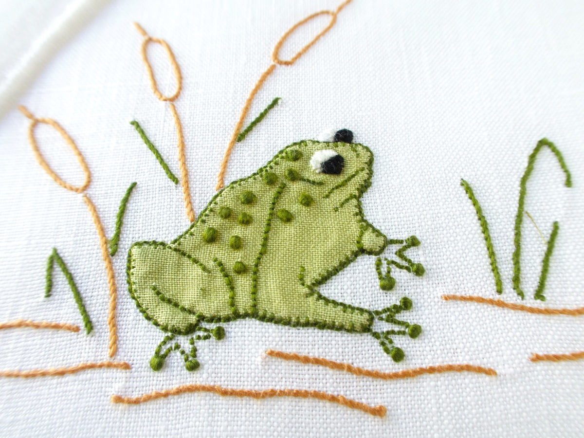 Close-up of a vintage Madeira cocktail napkin, white linen with scalloped edges, appliqué green spotted frogs among cattails and reeds
