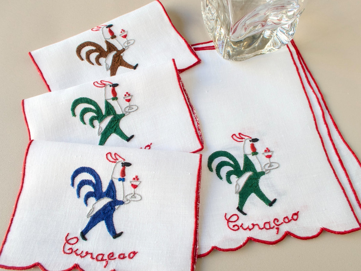 Stacked and folded vintage Madeira hand embroidered rooster cocktail napkins, white linen with red trim.