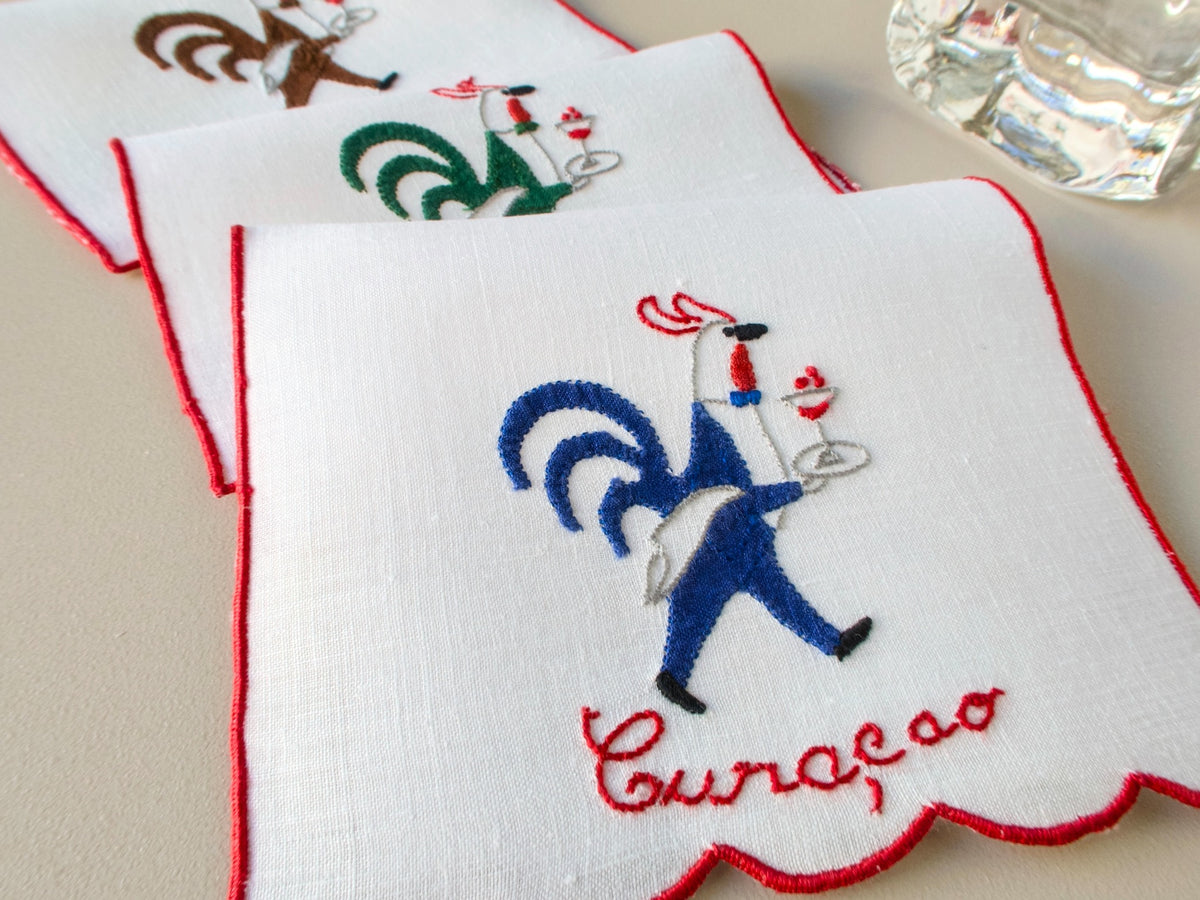 Vintage Madewira hand embroidered rooster cocktail napkin, white linen with red trim, placed on a table.