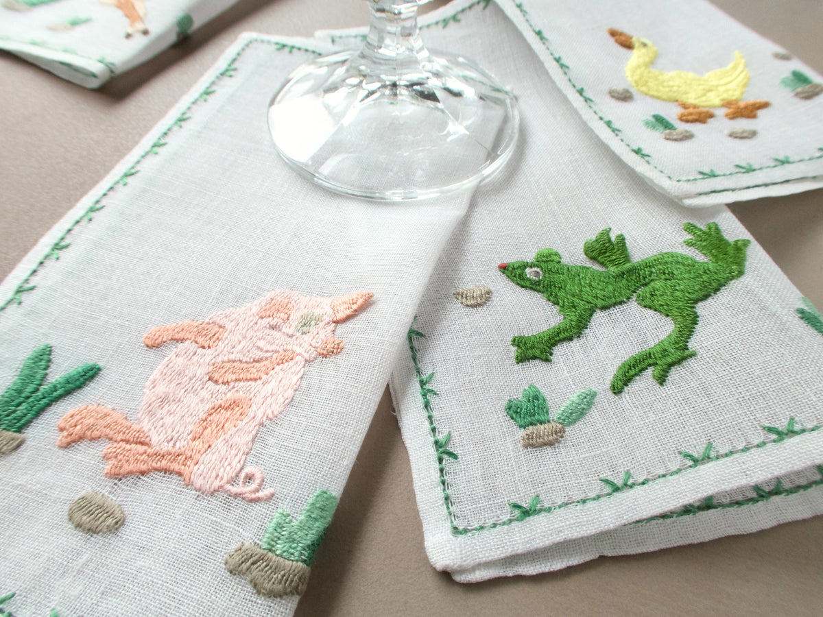 Close-up of Rapisardi animal cocktail napkins with embroidered pig and frog under a wine glass.