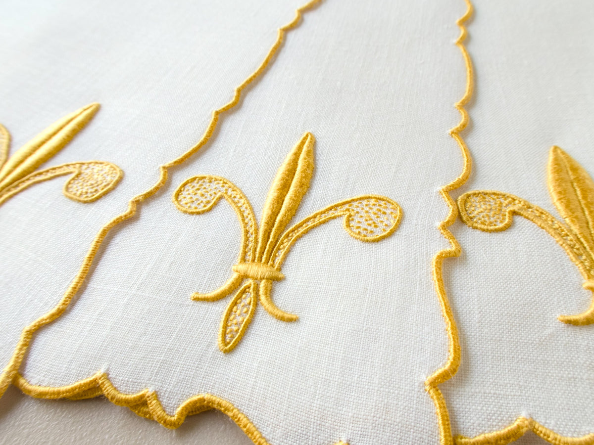  Detail of vintage Madeira cocktail napkin showing multiple gold fleur-de-lis motifs along scalloped edge, white linen​​​​​​​​​​​​​​​​