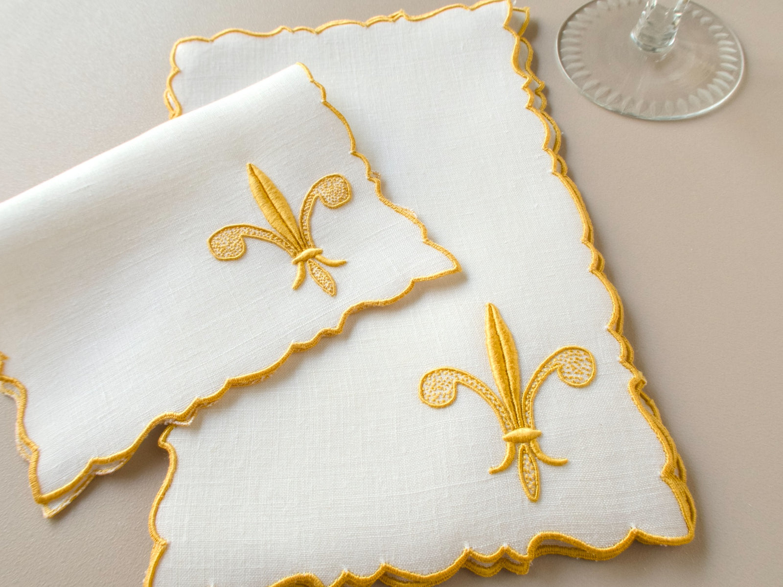Two vintage Madeira white linen cocktail napkins, raised gold fleur-de-lis embroidery, scalloped edges, styled with cocktail glass