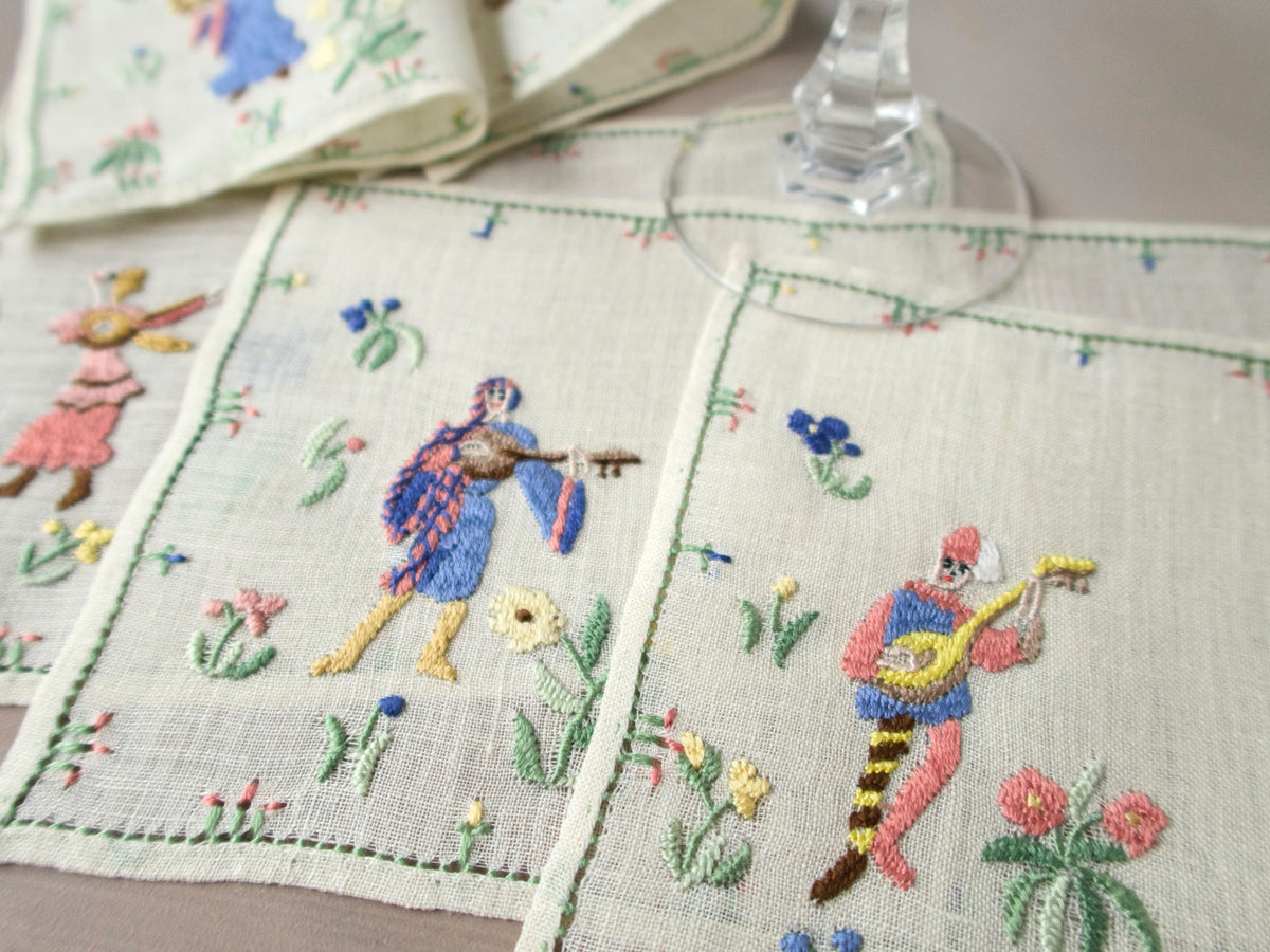Close-up of Rapisardi medieval musician cocktail napkins with two figures and floral embroidery under a glass stem.
