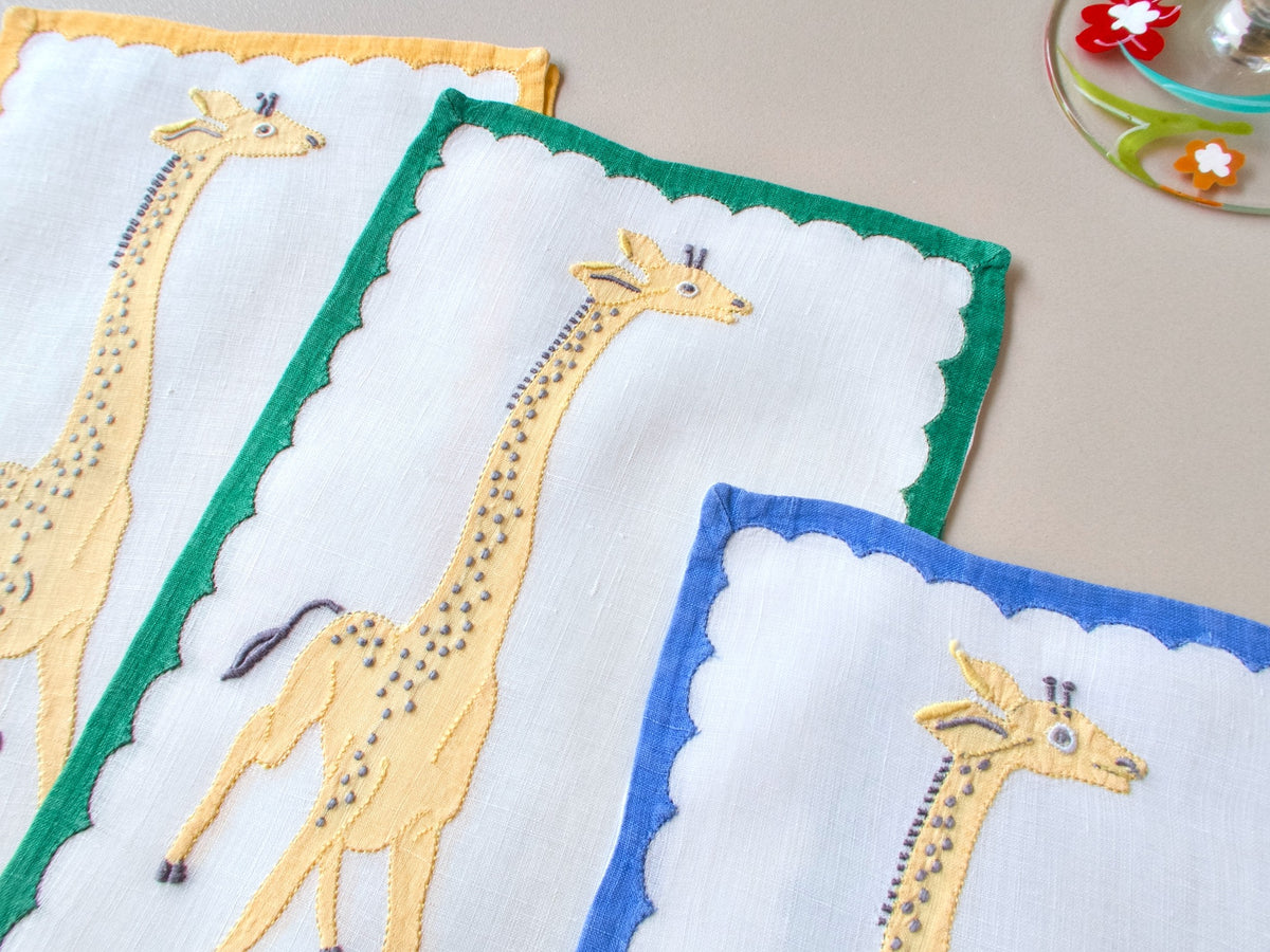 Three vintage Madeira cocktail napkins fanned out, white linen with green, yellow and blue borders, appliqué and embroidered giraffes in golden tones