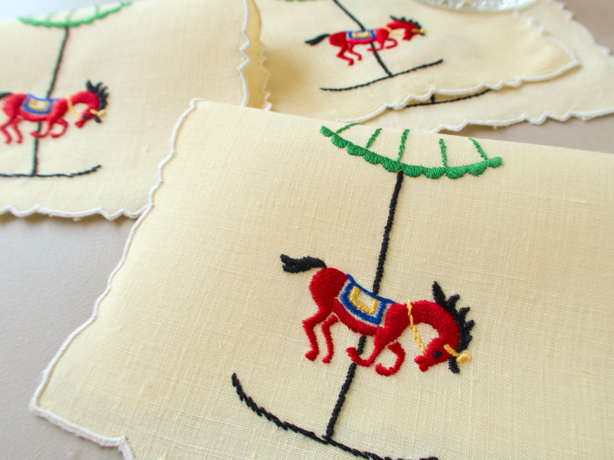 Close-up of a vintage Madeira cocktail napkin, yellow linen with scalloped edges, embroidered red carousel horse with blue and gold saddle on a black pole