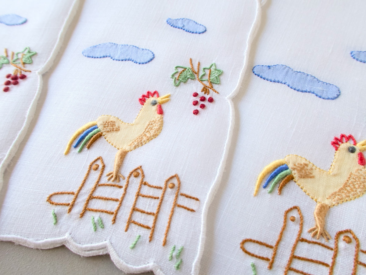 Close-up of vintage Madeira cocktail napkin, white linen with scalloped edges, embroidered rooster on fence post with rainbow tail feathers, red grape cluster and blue clouds