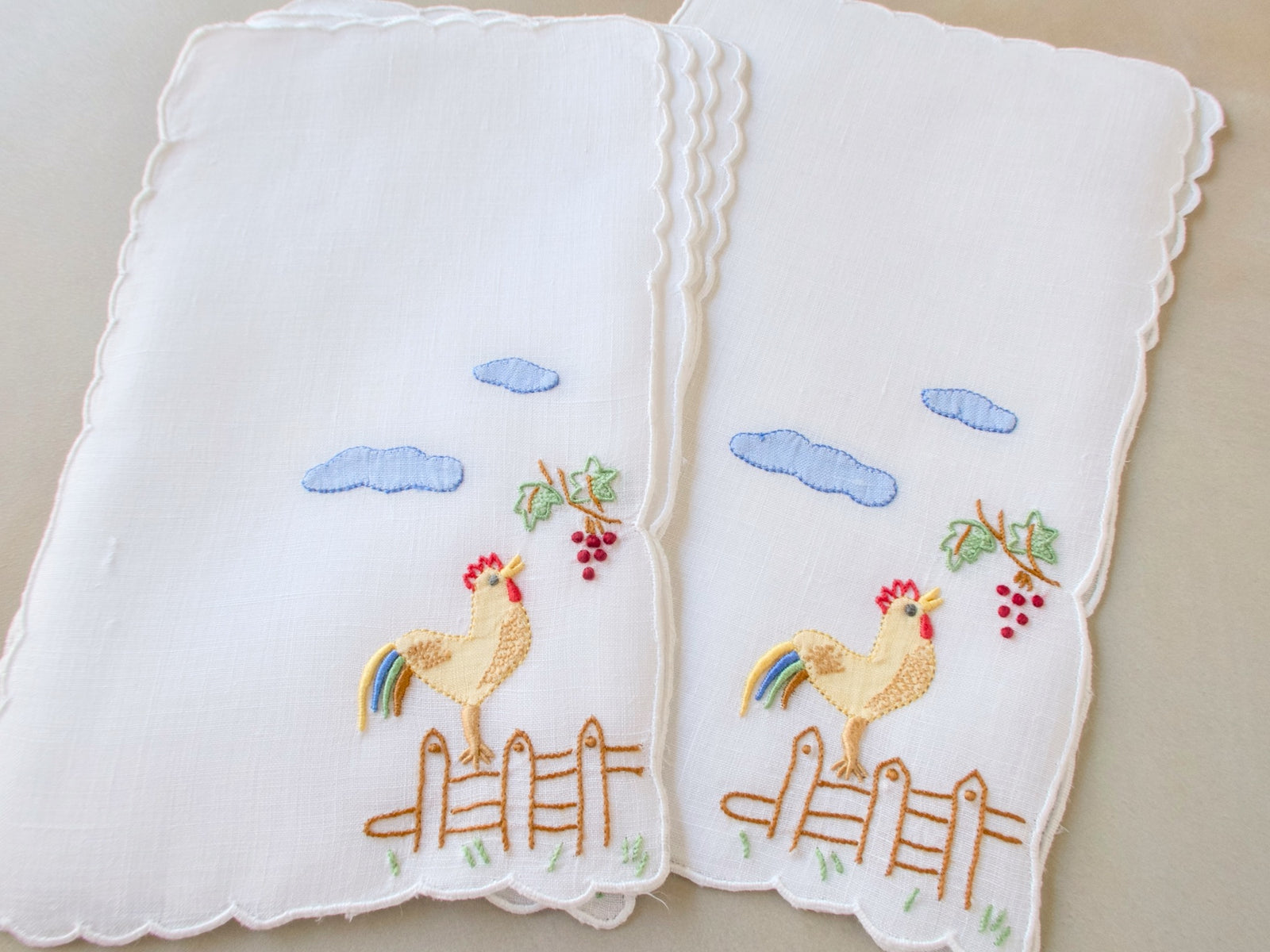 Stacked vintage Madeira cocktail napkins, white linen with scalloped edges, embroidered rooster on fence post with rainbow tail feathers, red grape cluster and blue clouds.