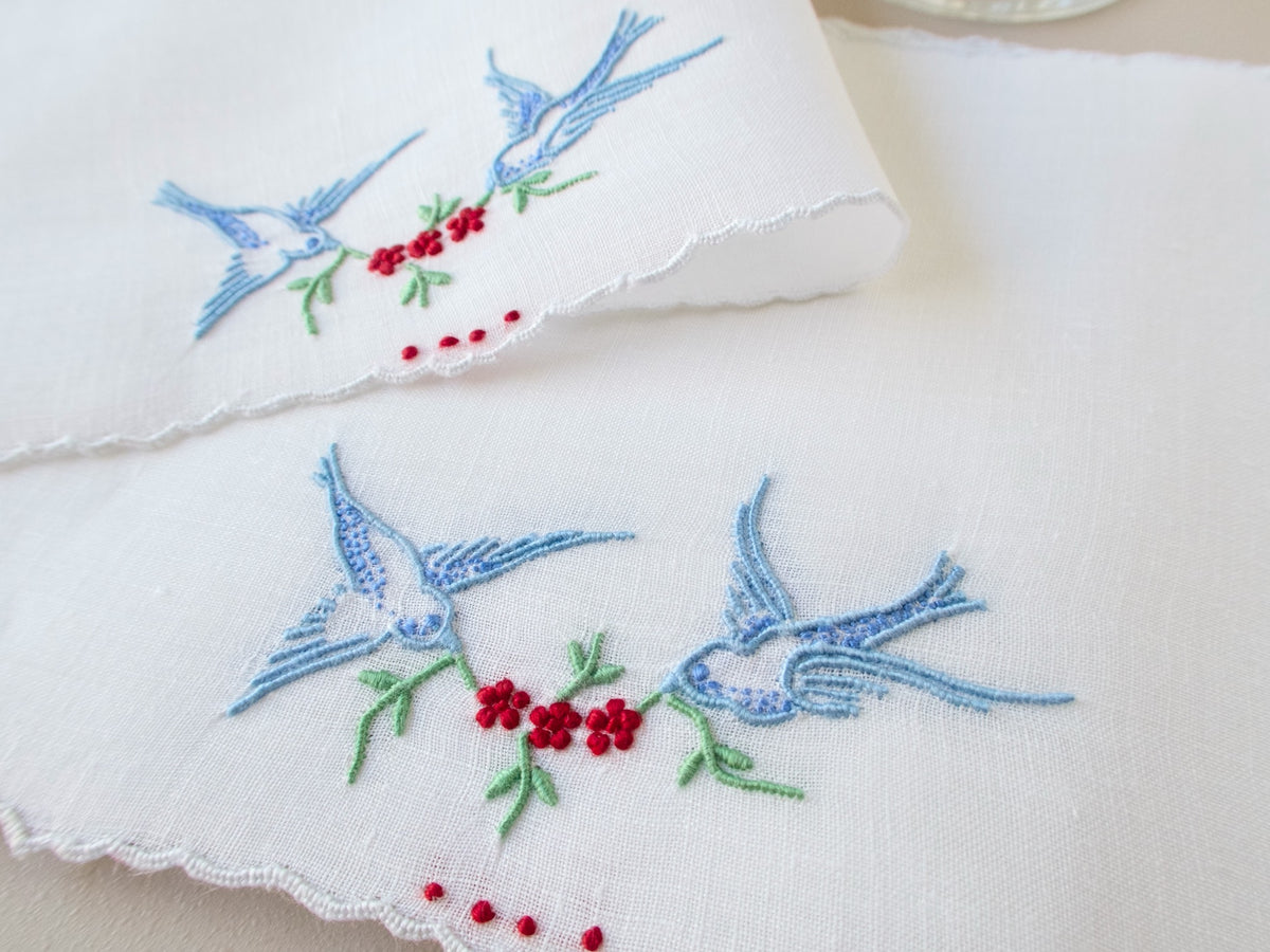 Close-up of vintage Madeira cocktail napkins, white Irish linen with scalloped edges, bluebirds in flight carrying a garland of red flowers.