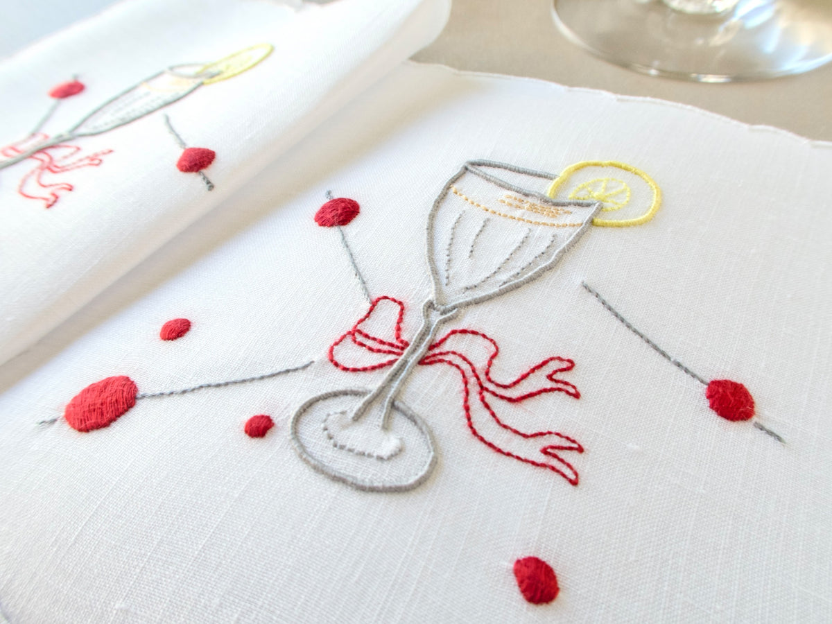 Close-up of a vintage Madeira cocktail napkin, white Irish linen with scalloped edges, embroidered cocktail glass with red bow, lemon slice and cherry picks​​​​​​​​​​​​​​​​