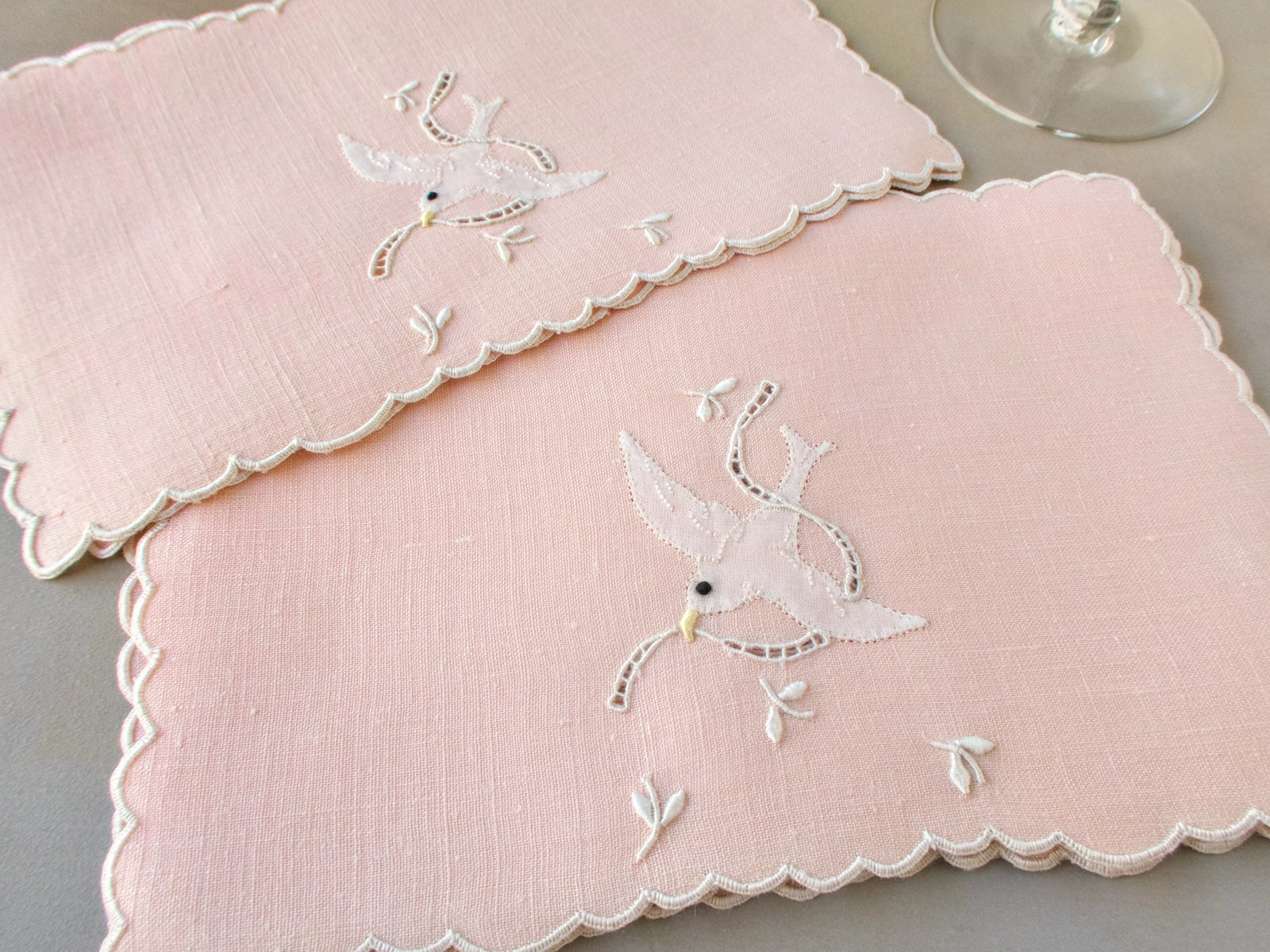Closeup of vintage Madeira cocktail napkins, blush pink linen with white scalloped edges, appliqué dove in flight with falling blossoms and cutwork ribbon